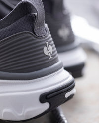 Close-up of a dark gray and white athletic shoe showing the textured side panel, heel logo, and chunky sole.