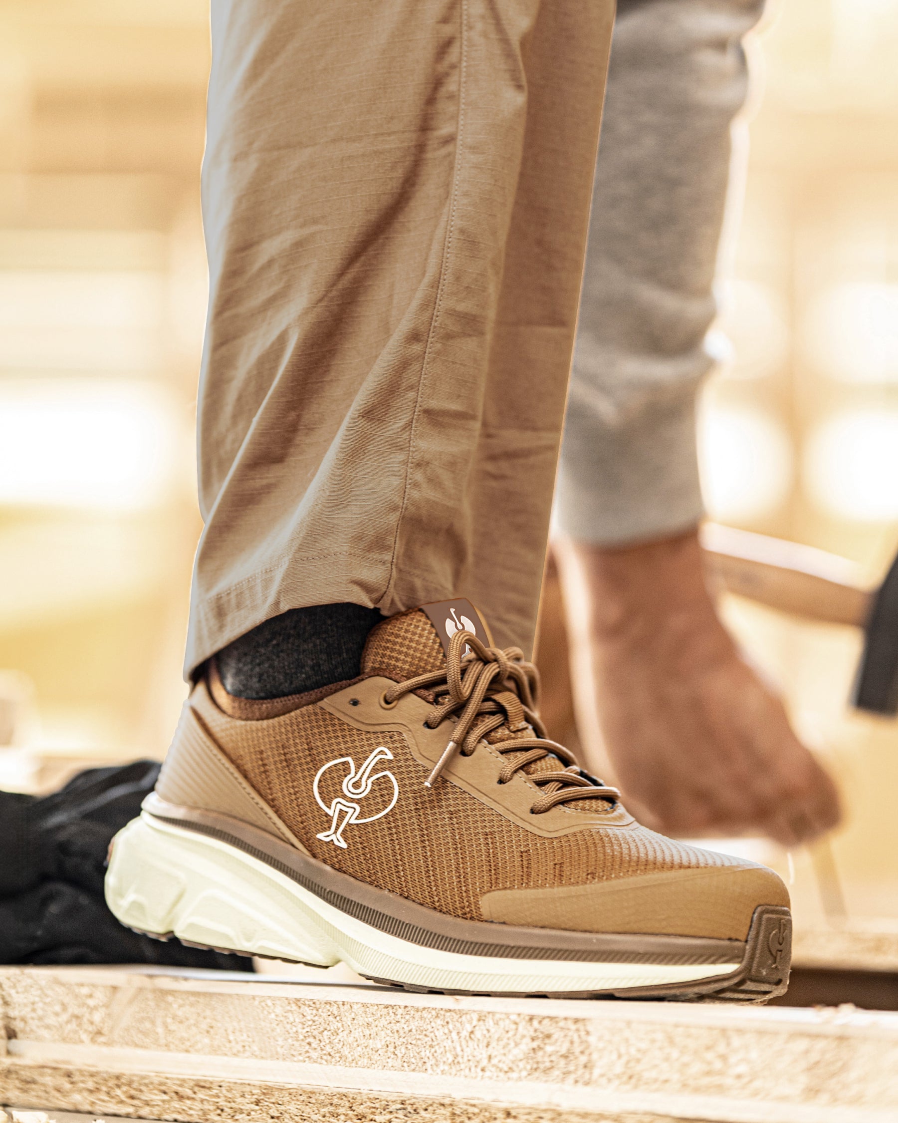 A close-up of a person's foot wearing a tan and brown athletic shoe with a thick white sole, standing on a stone surface. The person is also wearing khaki pants and a gray sweatshirt, with a blurred outdoor background.