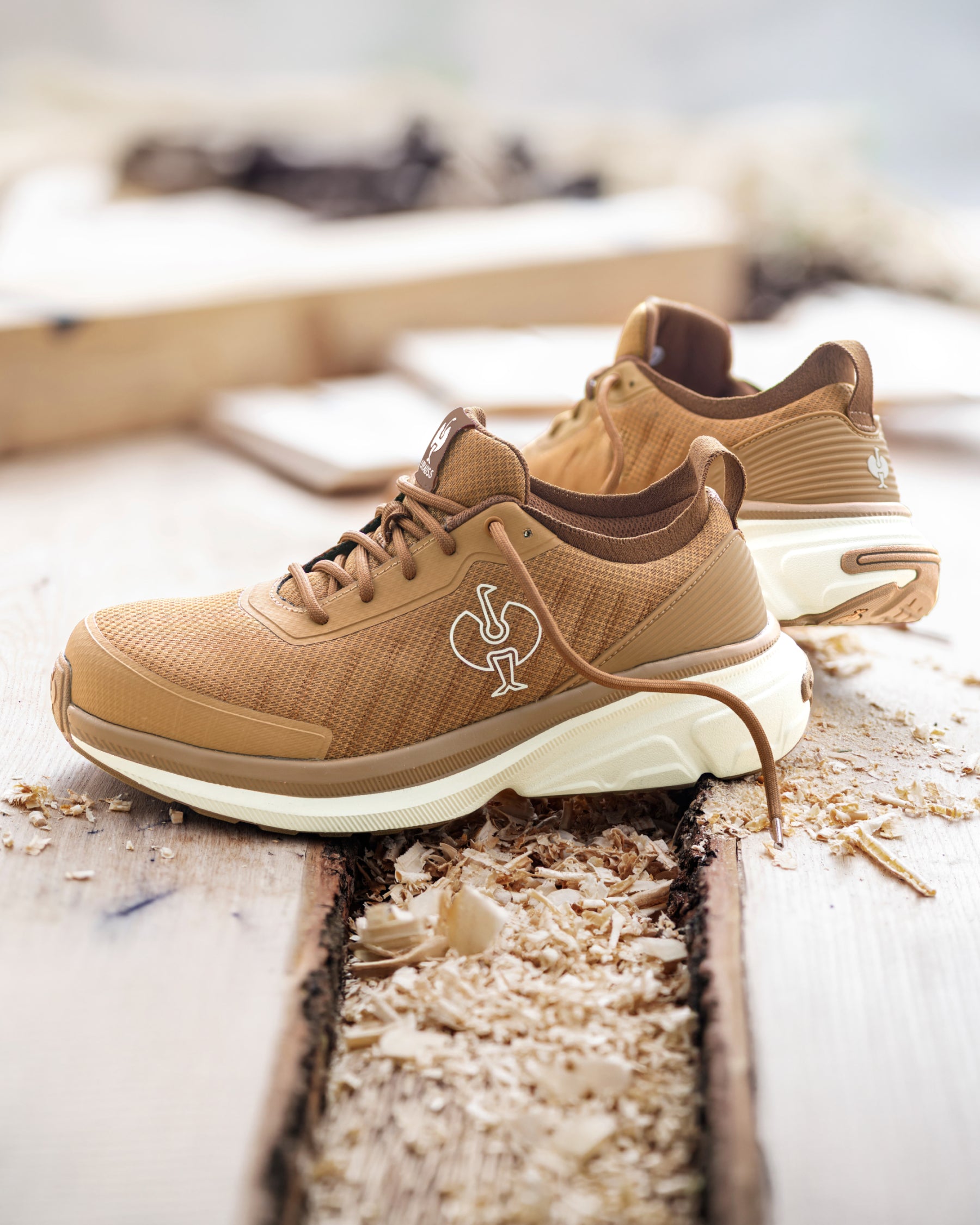A pair of tan and brown athletic sneakers with a white sole, styled on a rustic wooden surface with wood shavings. The shoe features a white rooster logo on the side.