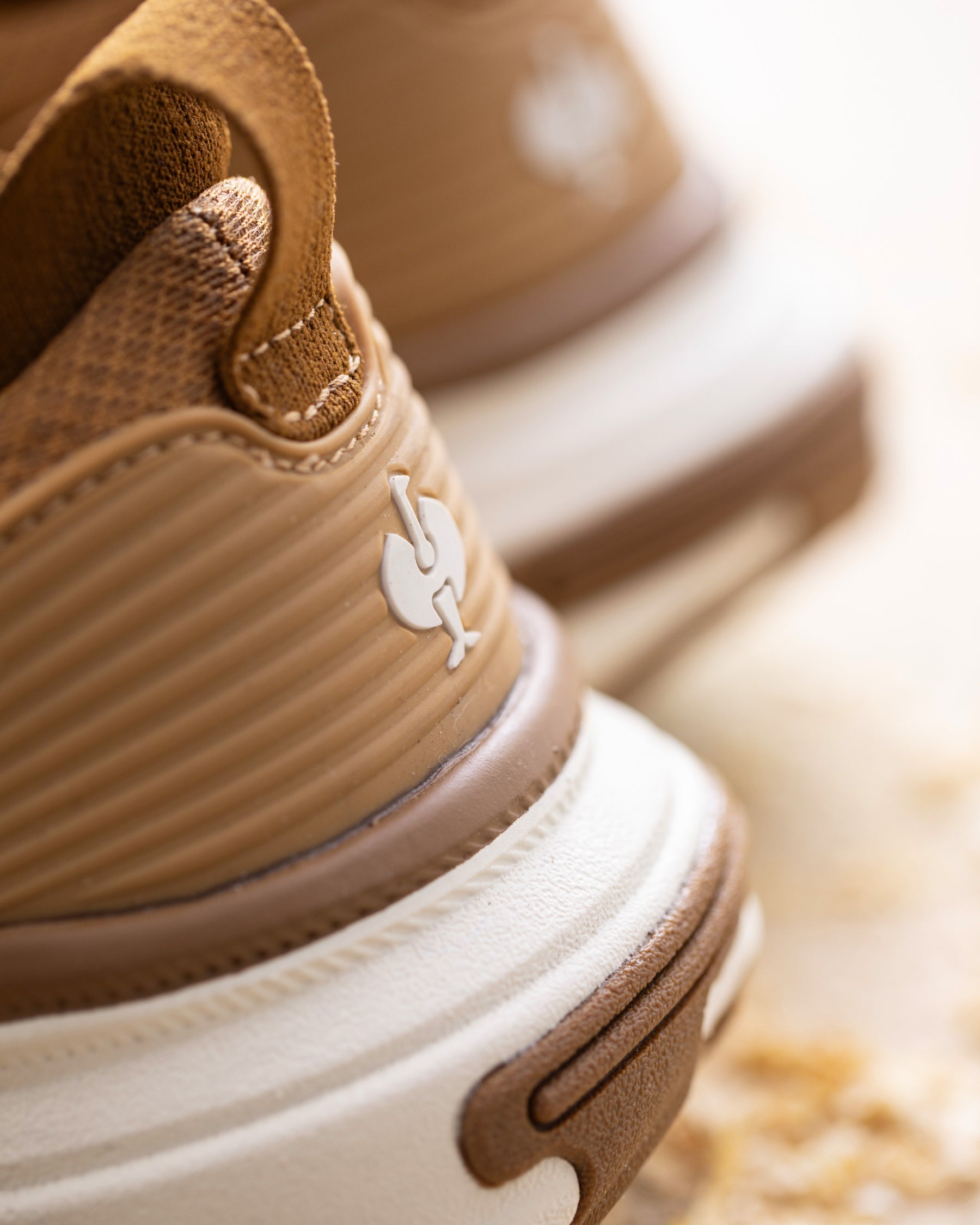 Close-up of a tan and brown sneaker showing the side profile, textured upper, pull tab, and thick white and brown sole, with a blurred second shoe in the background.