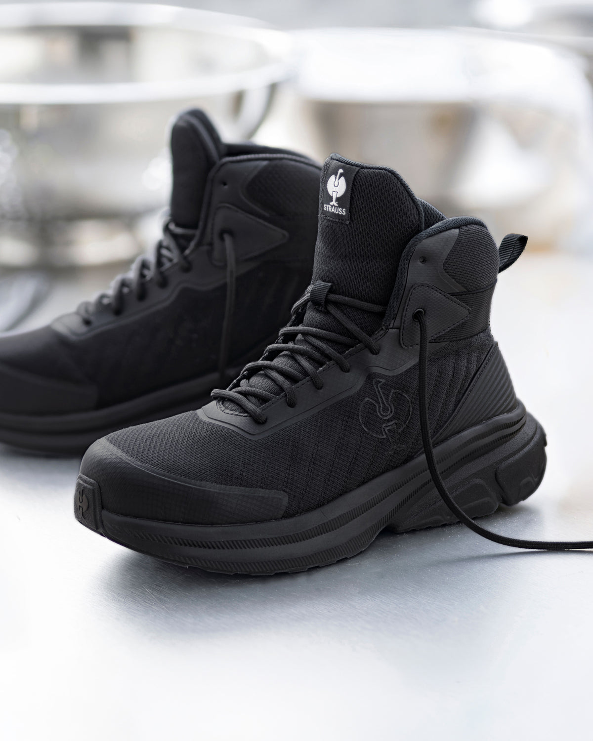 A pair of black high-top tactical boots with matching black laces and soles, displayed on a reflective light gray surface with a blurred studio background.