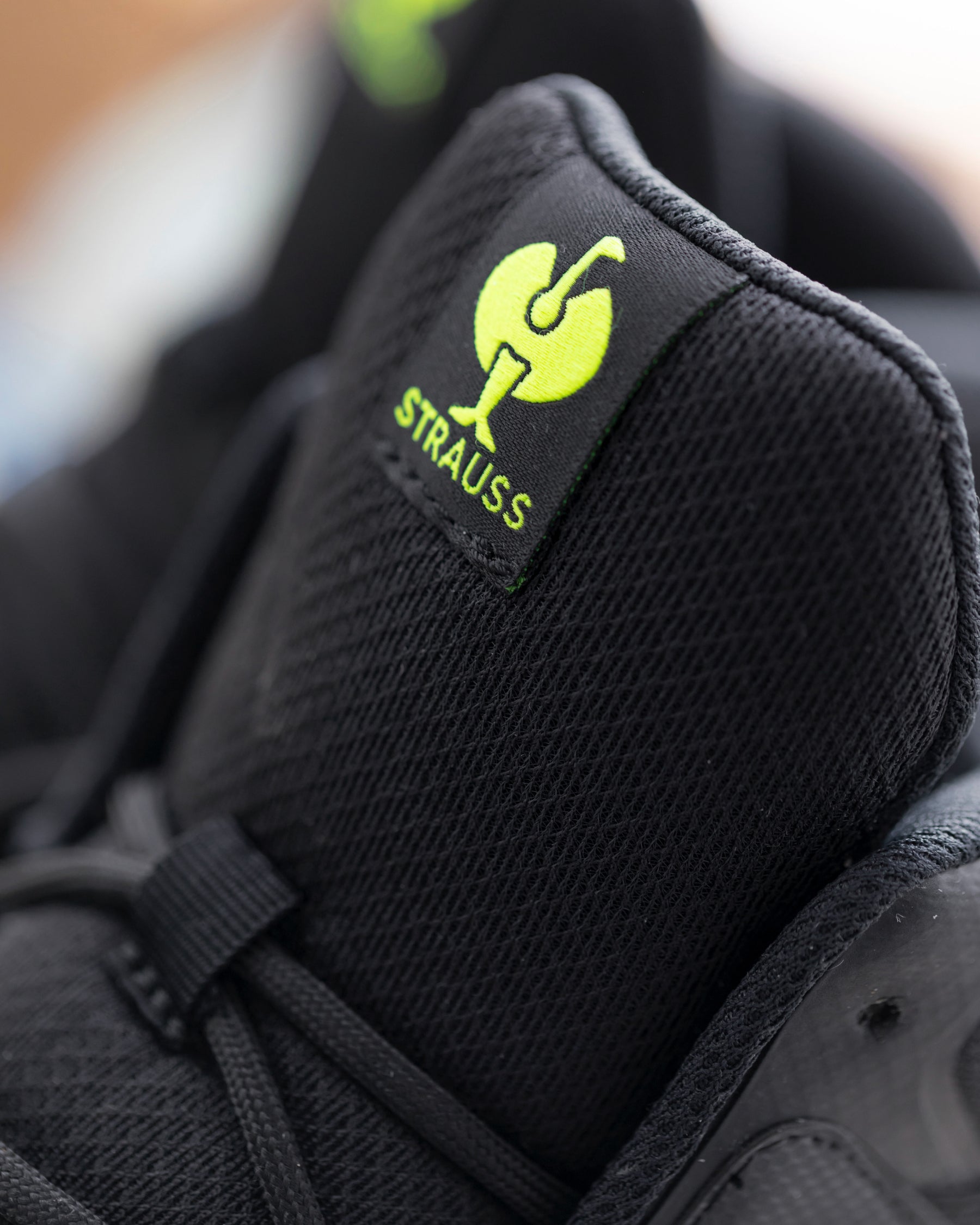 Close-up of a black mesh sneaker showing the tongue label with a bright yellow-green Strauss logo and text.