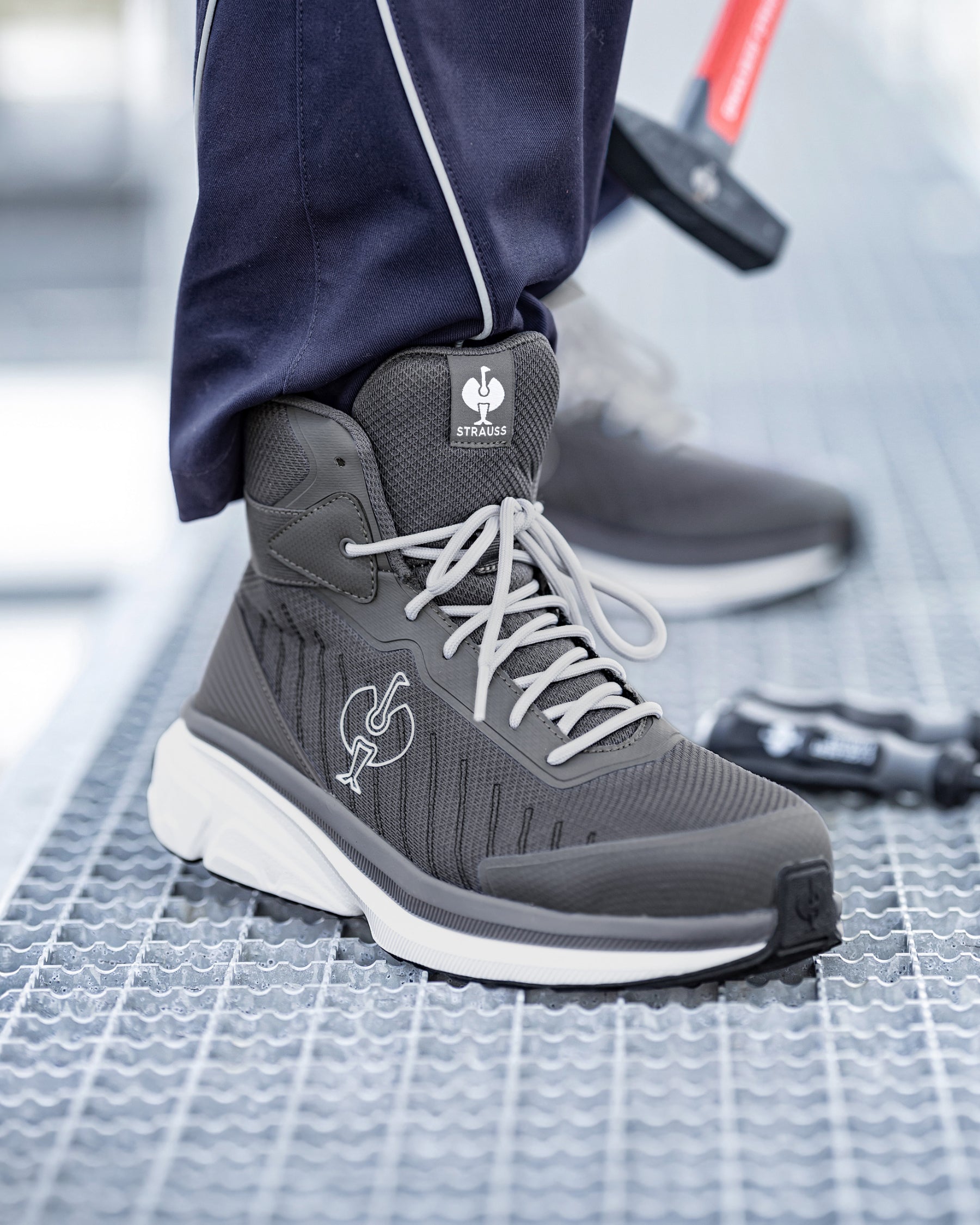 A close-up of a person wearing a dark gray and white high-top athletic boot with white laces, standing on a textured metal grate. The brand logo on the tongue and side reads 'SAS'.