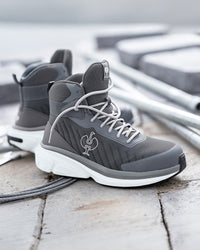 A pair of mid-top athletic sneakers in dark gray and white, featuring a textured mesh upper, white laces, and a white sole, displayed on a weathered wooden surface with a blurred industrial background.