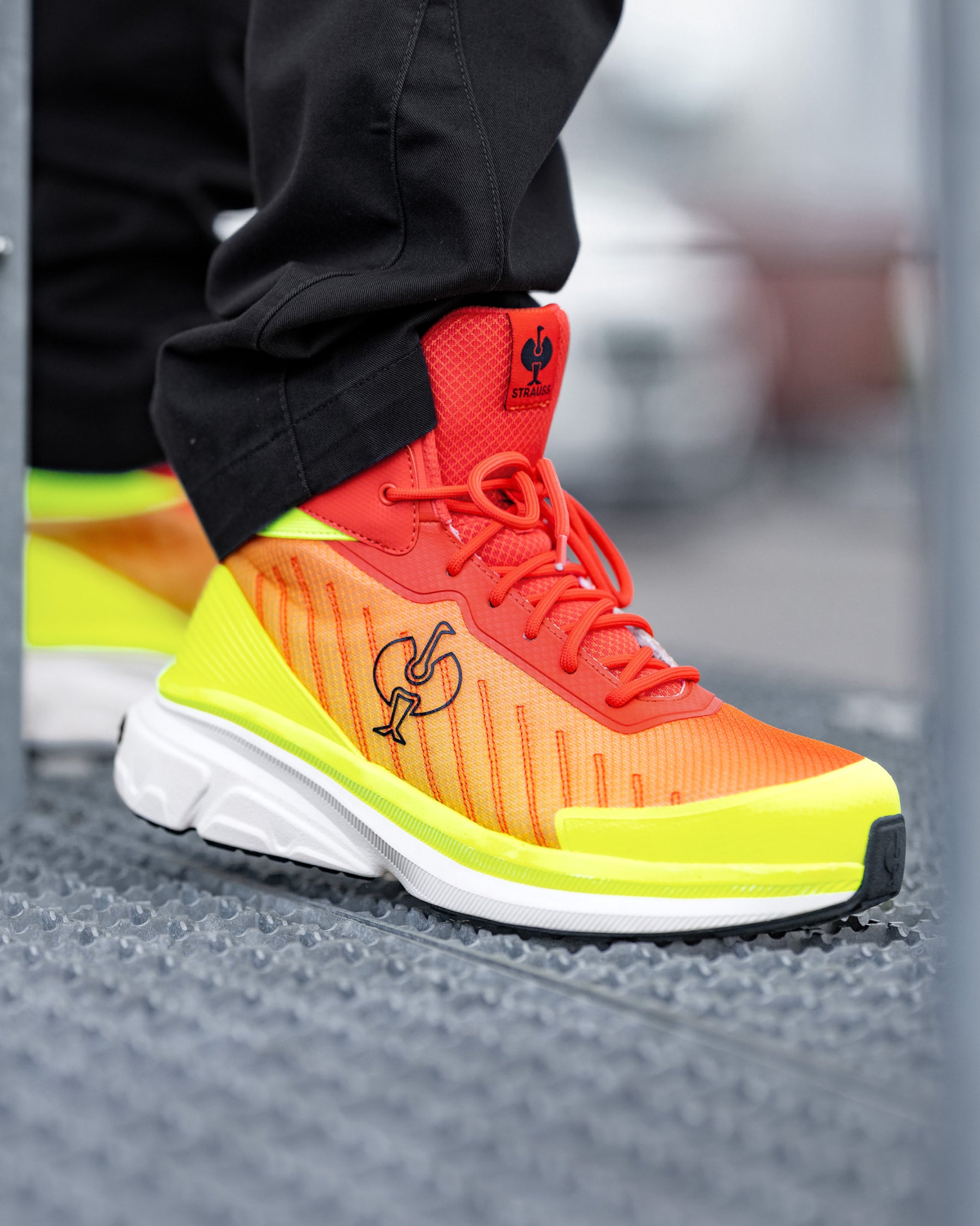 A close-up of a person's foot wearing a bright orange and neon yellow running shoe with black pants, standing on a textured gray metal grate.
