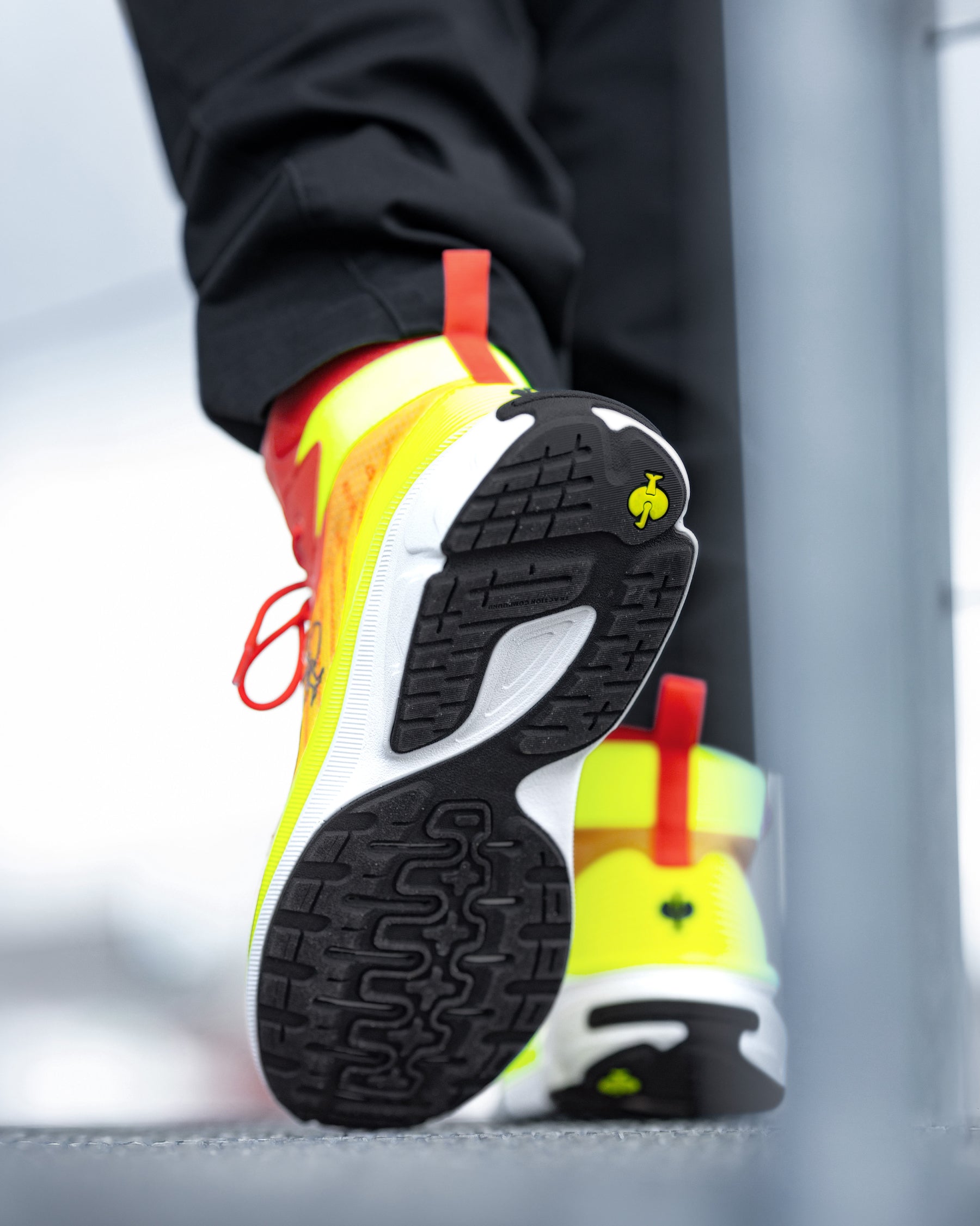 A close-up lifestyle shot of a person's foot wearing a bright neon yellow and white athletic shoe with a black treaded sole, standing on a gray surface.