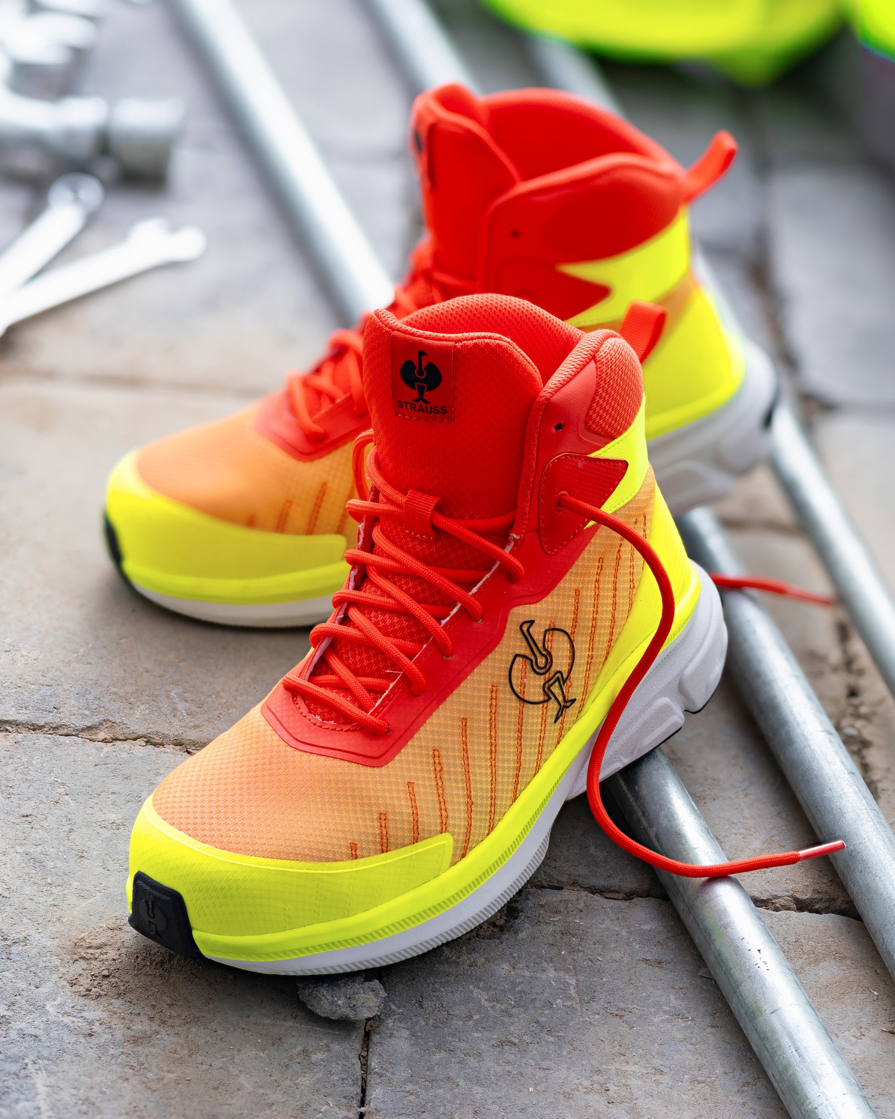 A pair of high-top athletic sneakers in a bright orange-to-yellow gradient with vivid red overlays, red laces, and a white midsole, photographed on a stone surface next to metal pipes.