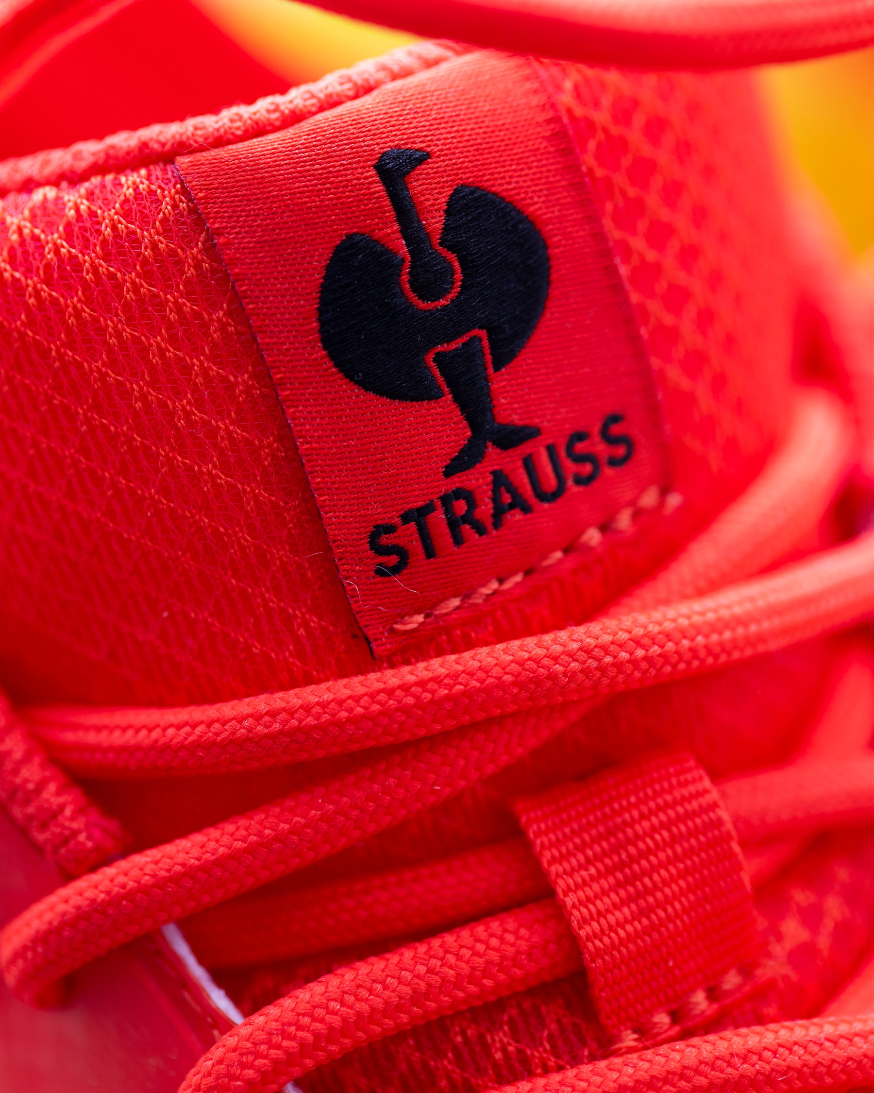 Close-up of a bright red sneaker showing the tongue label with a black Schoenstaettchen logo and the word 'STRAUSS' in black, with red laces and mesh texture.