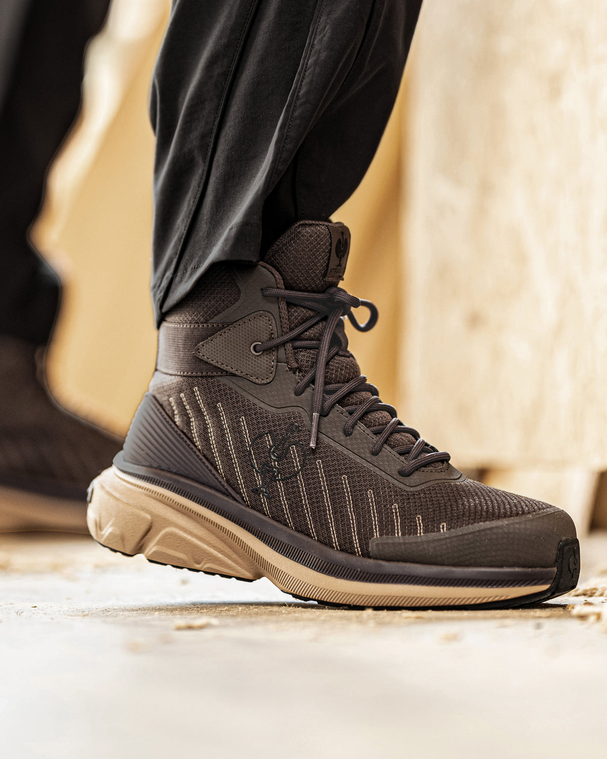A close-up of a person's foot wearing a dark brown and black mid-top athletic shoe with a beige sole, paired with black pants, standing on a concrete surface.