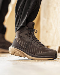 A close-up of a person's foot wearing a dark brown and black mid-top athletic shoe with a beige sole, paired with black pants, standing on a concrete surface.