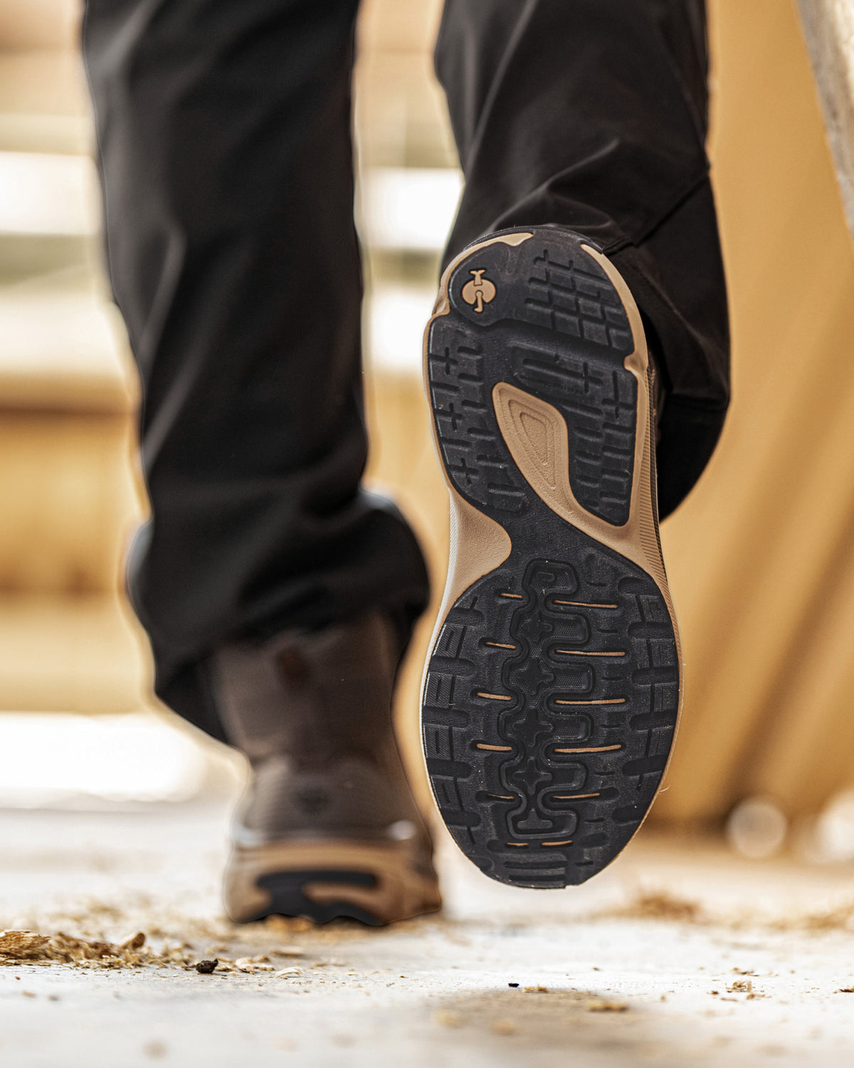 Close-up of a person's foot wearing black hiking boots and black pants, lifting the heel to show the textured black and tan outsole of a hiking shoe.