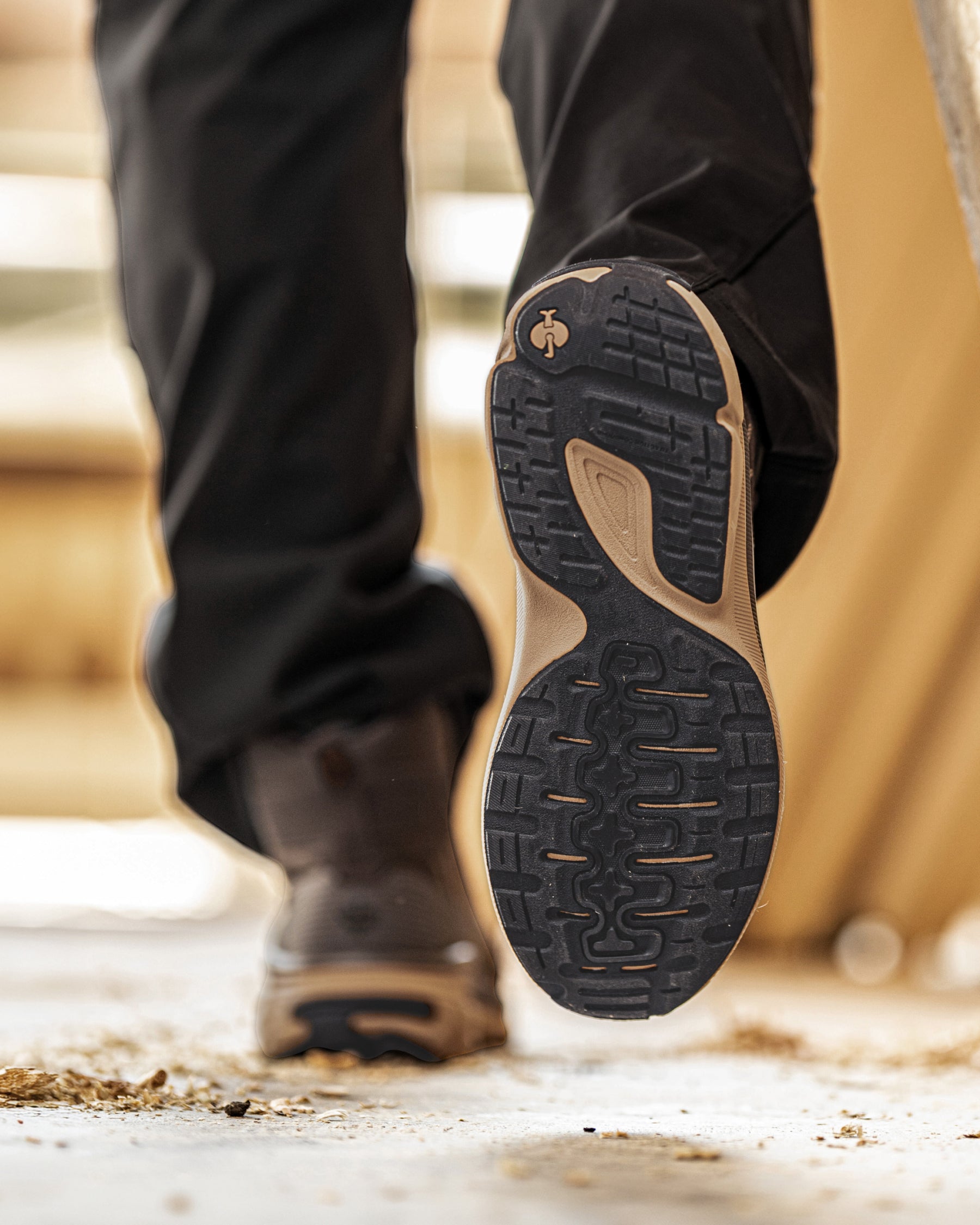 Close-up of a person's foot wearing black hiking boots and black pants, lifting the heel to show the textured black and tan outsole of a hiking shoe.