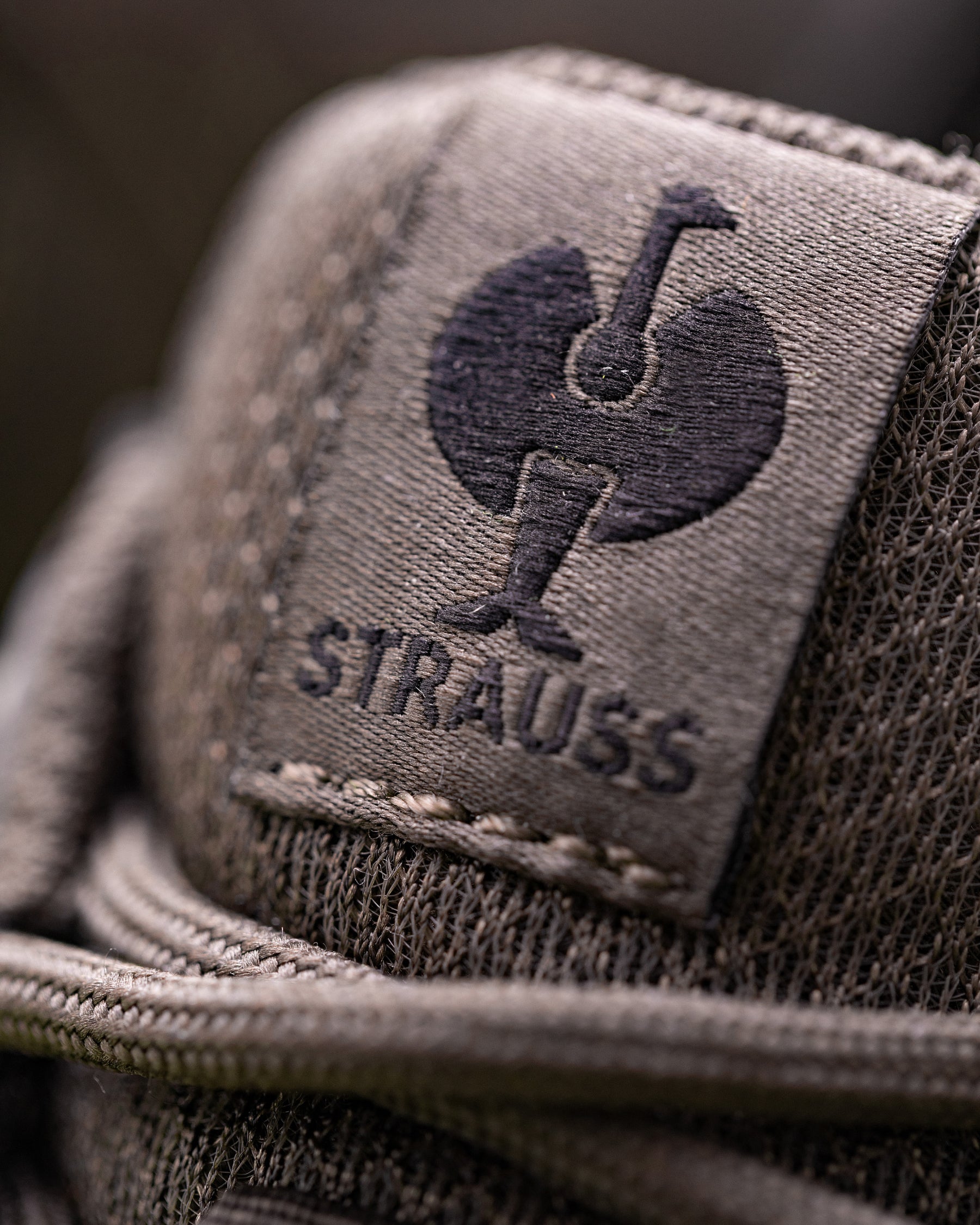 Close-up of a textured taupe woven fabric patch with a dark embroidered rooster logo and the word 'Strauss' beneath it.