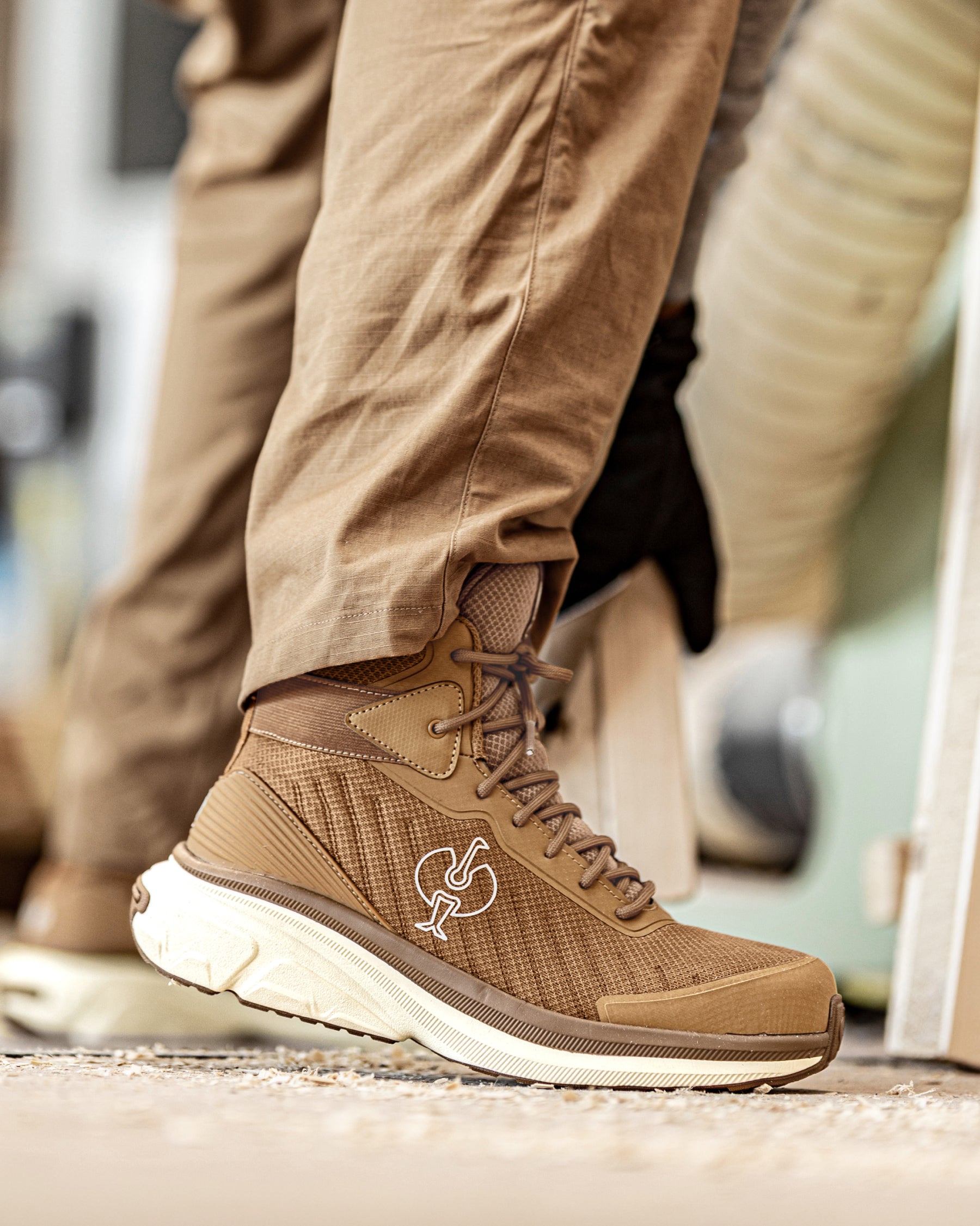 A close-up of a person's foot wearing a tan and brown mid-top athletic shoe with a white sole, paired with khaki pants.