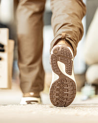 A close-up of a person walking, showing the textured brown and beige outsole of a sneaker in sharp focus, with tan pants and a blurred indoor background.