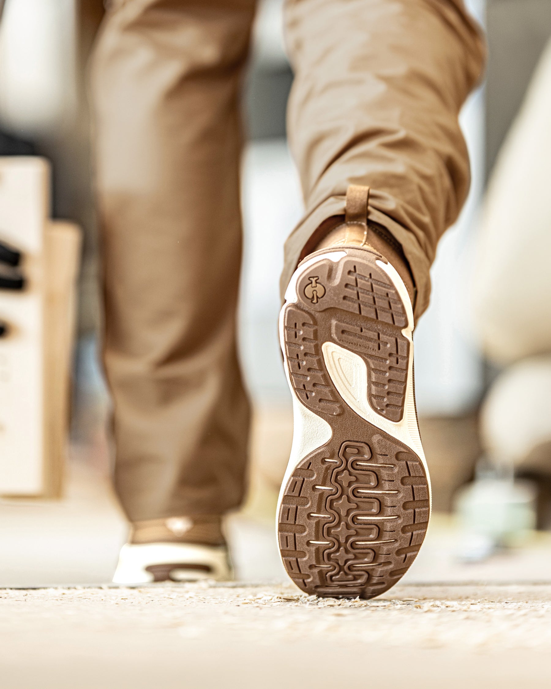 A close-up of a person walking, showing the textured brown and beige outsole of a sneaker in sharp focus, with tan pants and a blurred indoor background.