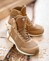A pair of tan high-top work boots with white midsoles, styled on a wooden surface with wood shavings. The boots feature a textured mesh upper, lace-up front, and a small logo patch on the tongue.