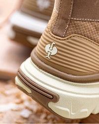 Close-up of a brown and beige athletic shoe showing the heel area with a small white rooster logo on the side, set against a blurred outdoor background.