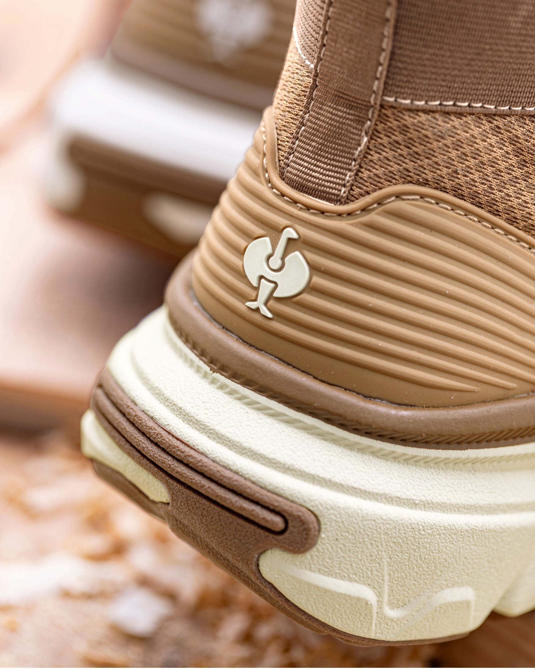 Close-up of a brown and beige athletic shoe showing the heel area with a small white rooster logo on the side, set against a blurred outdoor background.