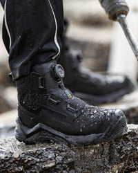 A close-up of a black waterproof work boot with a dial closure, standing on wet concrete next to a person's legs wearing black pants, with a power tool visible in the blurred background.