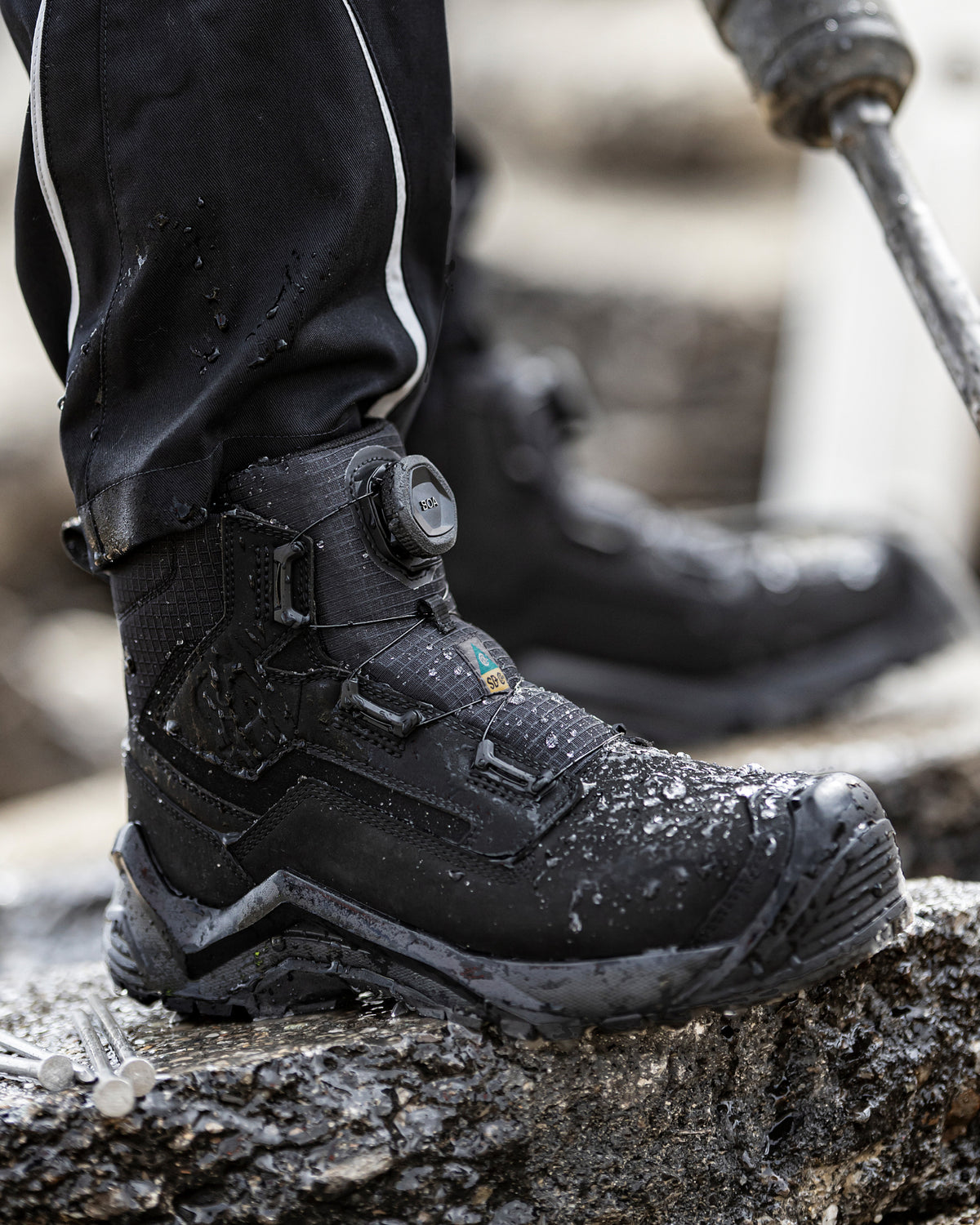 A close-up of a black waterproof work boot with a dial closure, standing on wet concrete next to a person's legs wearing black pants, with a power tool visible in the blurred background.