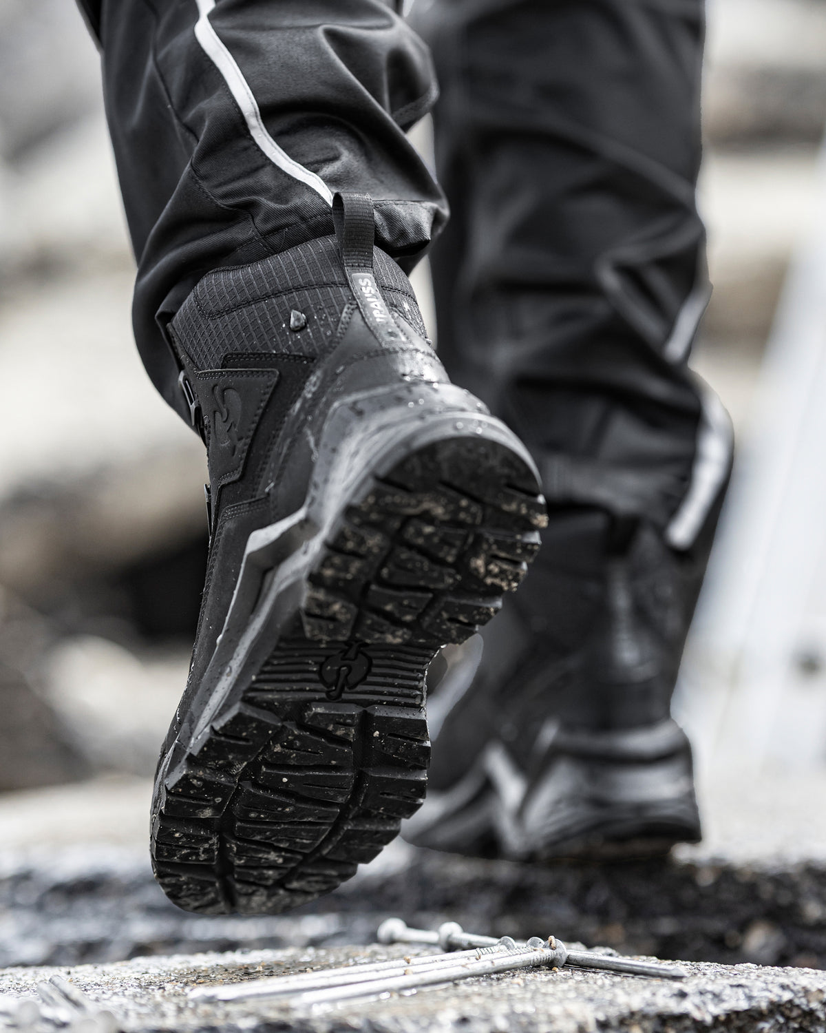 Close-up of a person wearing black technical riding boots and black pants with white piping, stepping on a rough outdoor surface. The boot on the left is lifted to show the treaded sole.