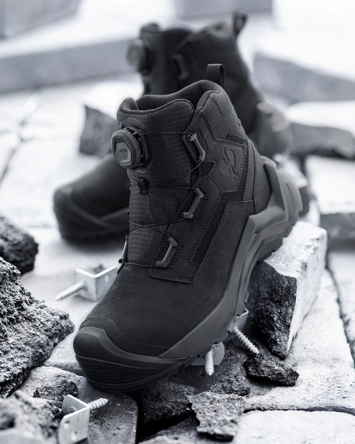 A pair of black tactical boots with a BOA dial closure system, displayed on broken concrete and metal fasteners in a monochrome, gritty setting.