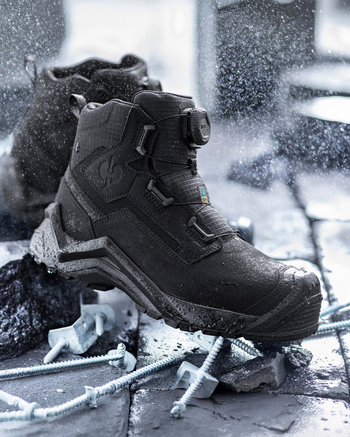 A black mid-cut work boot with a quick-release buckle strap, shown in a dynamic, snowy outdoor setting with screws on the ground.
