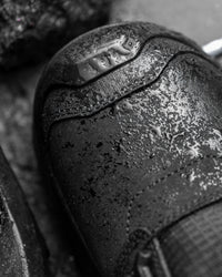 Close-up of a black leather shoe toe cap with water droplets and a metallic embossed logo, shown in a dark, moody monochrome photograph.
