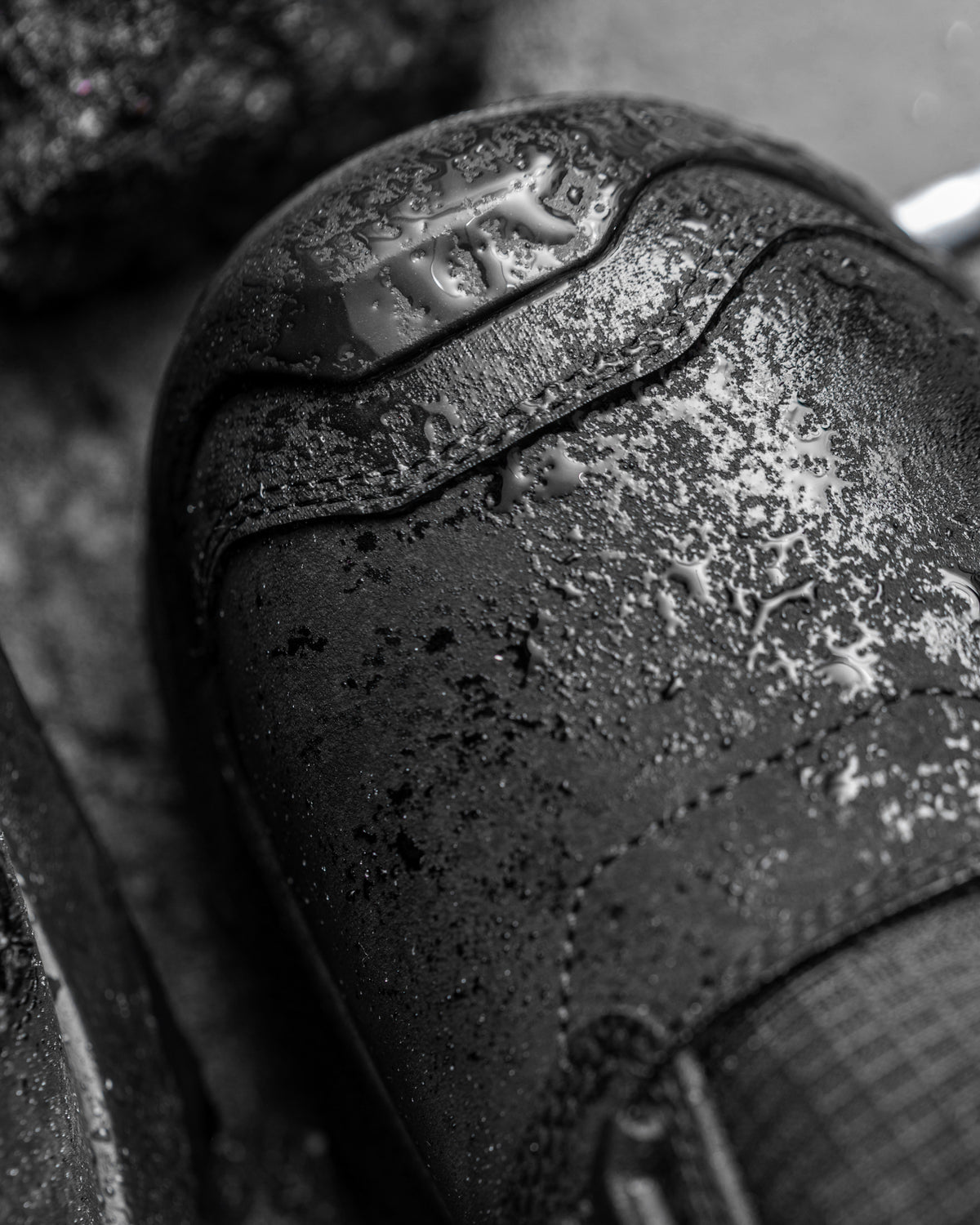 Close-up of a black leather shoe toe cap with water droplets and a metallic embossed logo, shown in a dark, moody monochrome photograph.