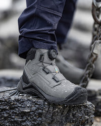 A close-up of a gray outdoor work boot with a dial closure, worn with dark navy pants and standing on a rough stone slab with a chain visible in the background.