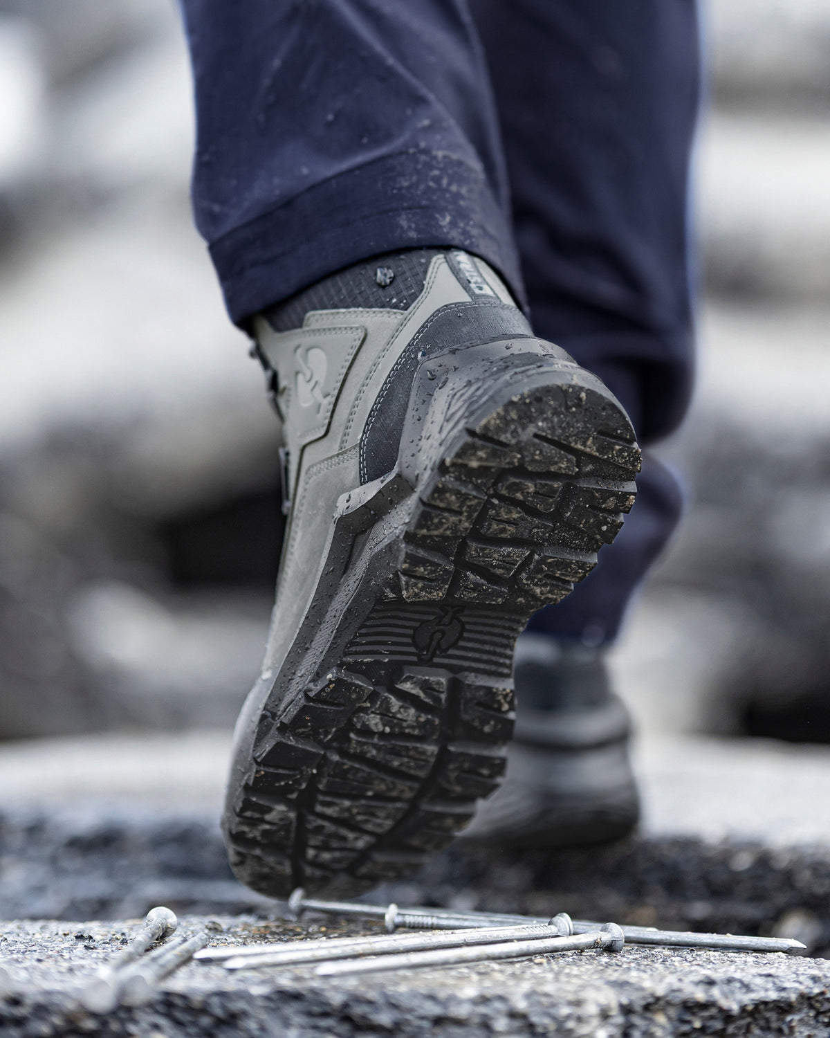 A close-up of a person's foot wearing a gray and black rugged work boot, stepping over metal nails on a rough concrete surface. The person's dark navy pants are also visible.