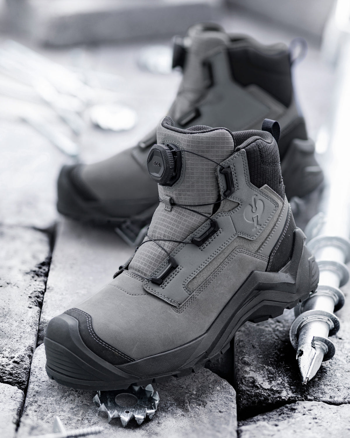 A pair of mid-cut tactical boots in a monochrome gray colorway, displayed on stone tiles with a metal pole as a prop.