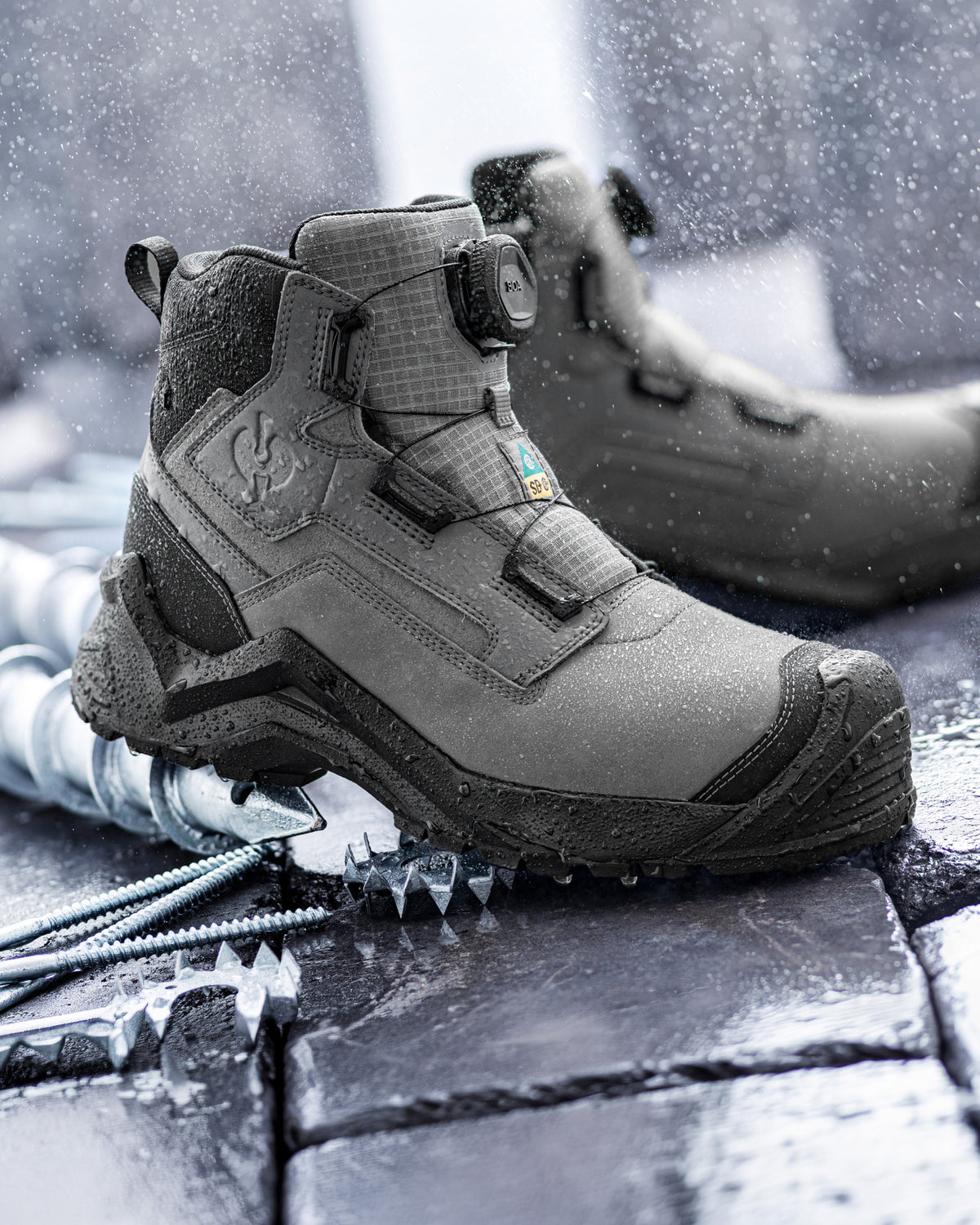 A pair of gray tactical winter boots with BOA dials, shown in a snowy, wet outdoor environment with ice picks on the ground.