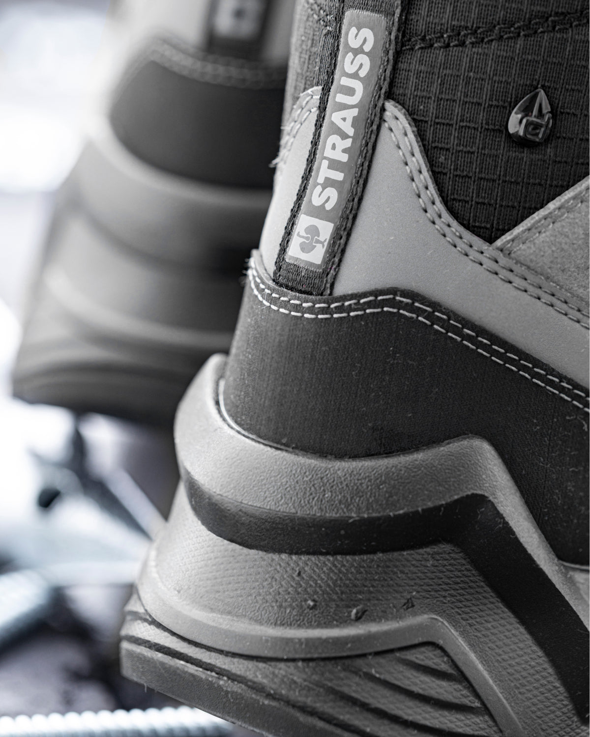 Close-up of a black and gray technical boot with a visible STRAUSS heel pull tab and textured synthetic overlays.
