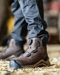 A close-up of a person wearing a brown dialux work boot with dark gray pants, standing on a bed of straw.