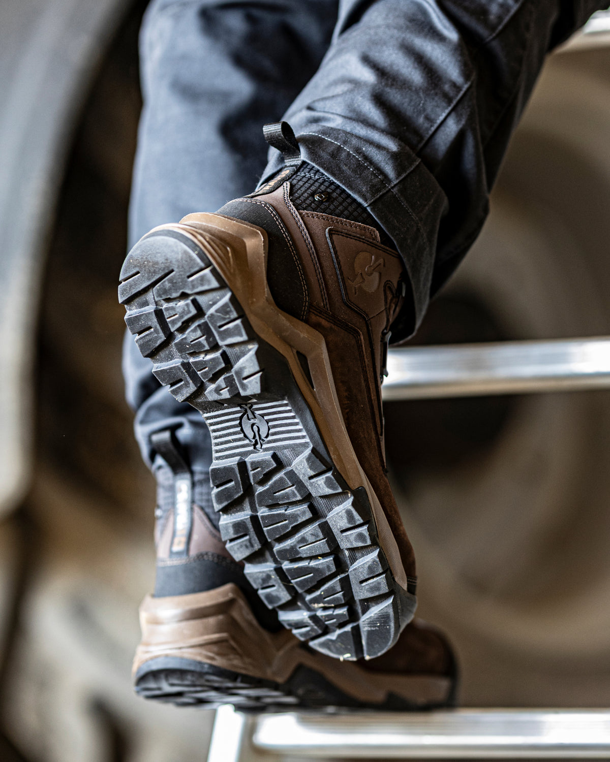 A close-up of a person's foot wearing a brown and black rugged work boot, showing the textured treaded outsole, with dark jeans and a metal ladder visible in the background.