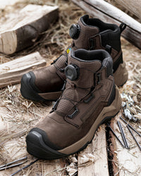 A pair of brown and black outdoor work boots with dial-up closures, displayed on a rustic background of wood and straw.