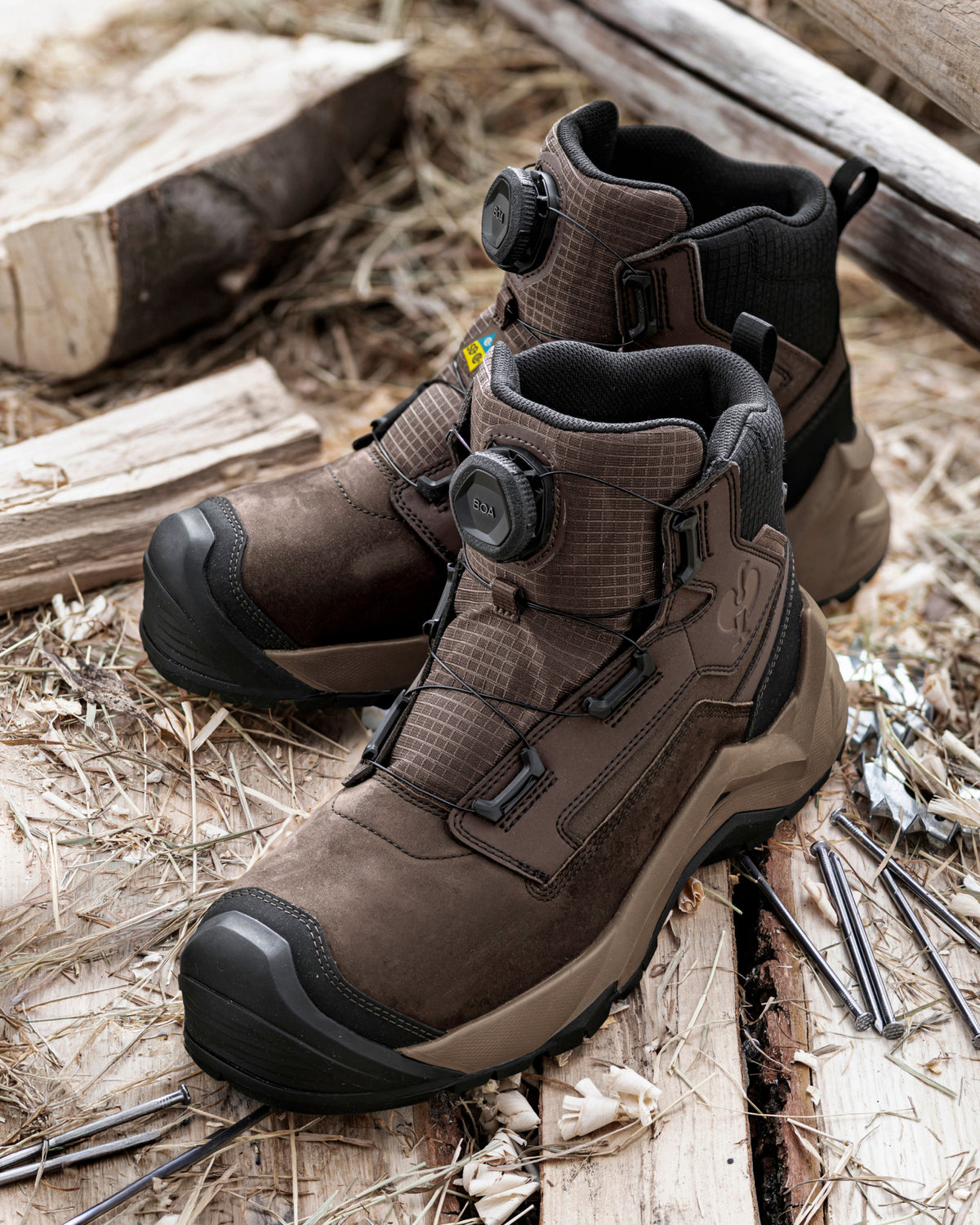 A pair of brown and black outdoor work boots with dial-up closures, displayed on a rustic background of wood and straw.