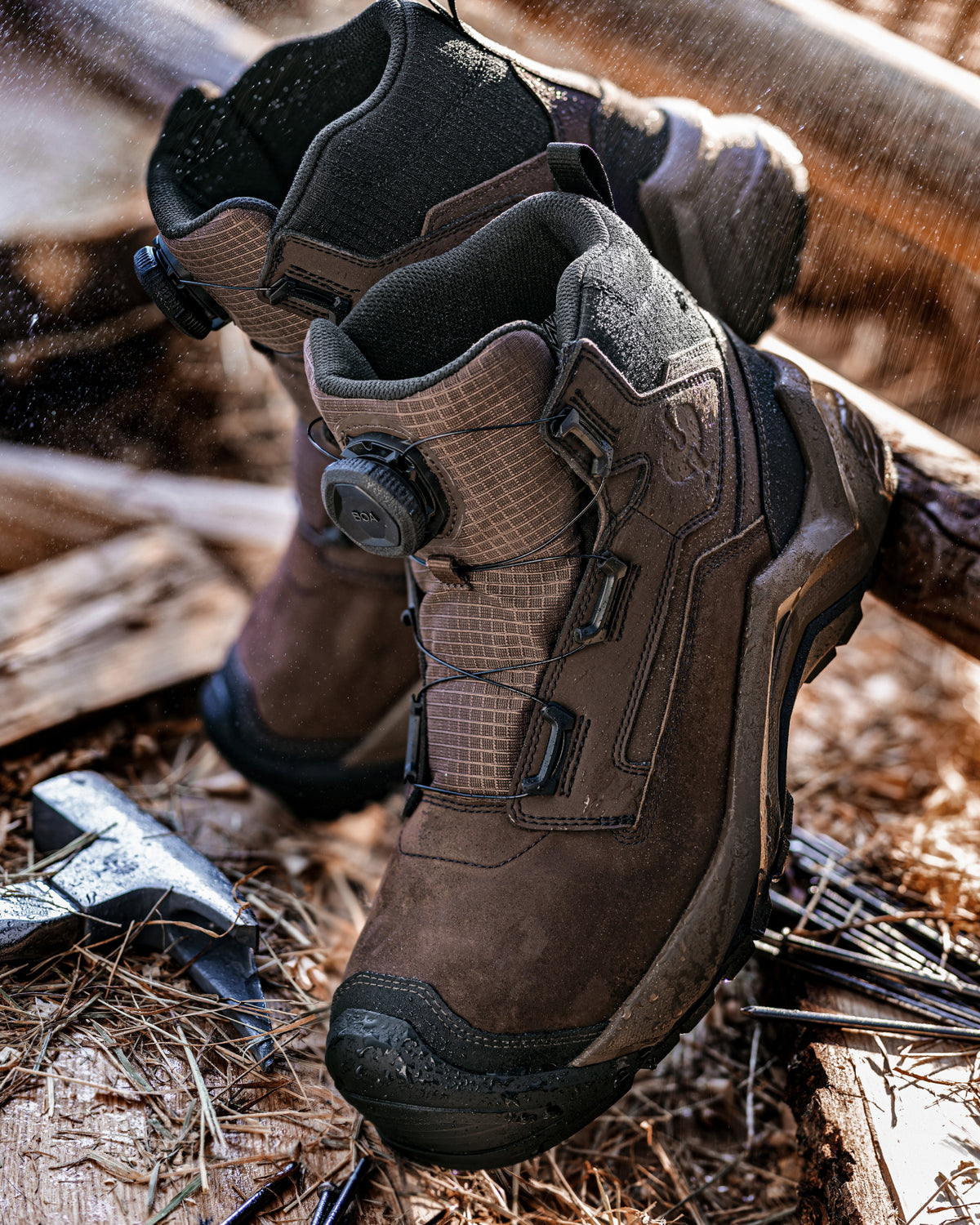 A pair of rugged brown and black tactical boots with BOA dials, shown outdoors on a bed of wood and pine needles.
