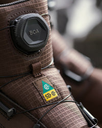 Close-up of a brown textured snowboard boot featuring a BOA dial closure and an SB C. G. brand patch.