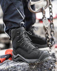 A close-up of a person wearing a black high-top work boot with gray laces, standing on a rough stone surface near heavy chains and industrial equipment.