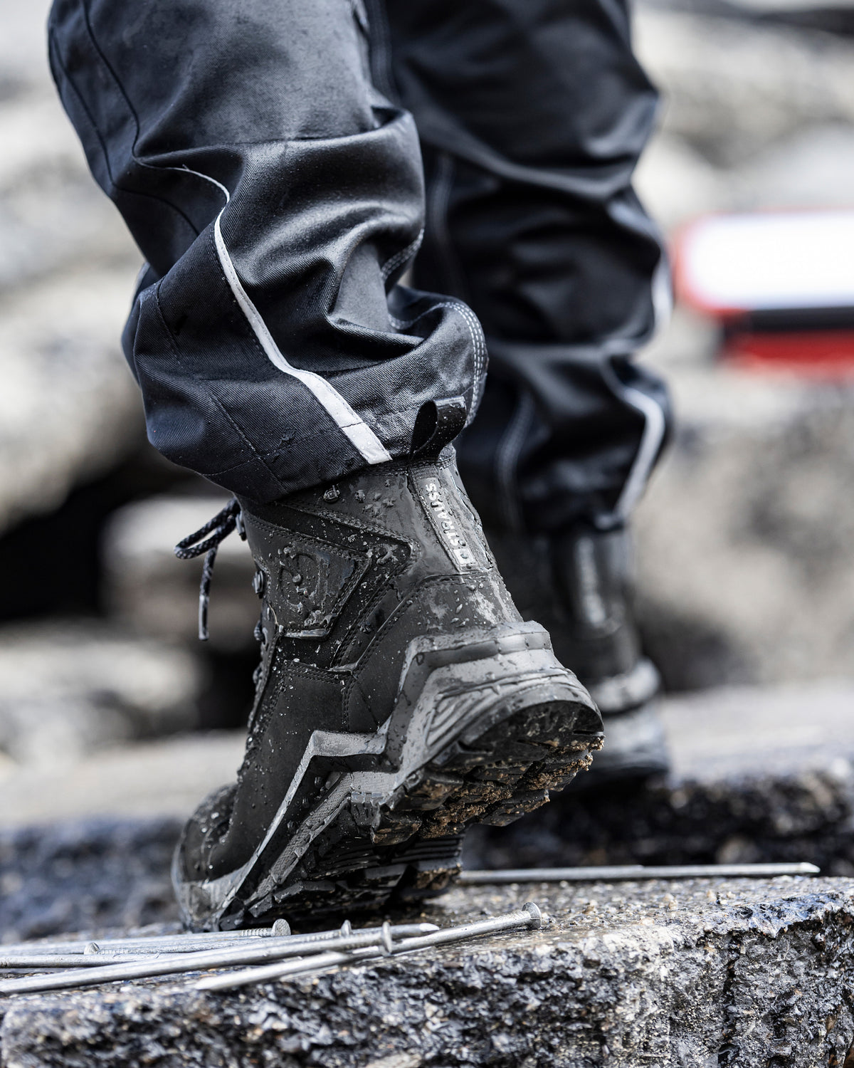 A close-up of a person's legs and feet wearing black waterproof pants and black high-top boots while stepping carefully over rough concrete rocks outdoors.