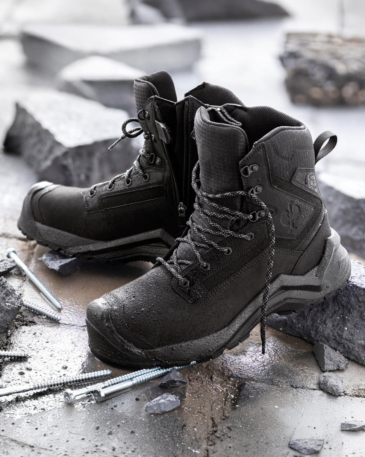 A pair of rugged black tactical combat boots with metal eyelets and laces, positioned on wet concrete amidst scattered rocks and loose screws.