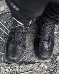 Black waterproof lace-up boots with water droplets on the surface, worn with black pants on a wet gray stone surface.