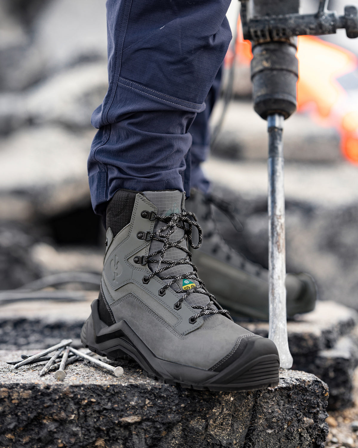 A close-up of a gray work boot with black laces, worn with navy pants on a rough concrete surface next to a jackhammer.