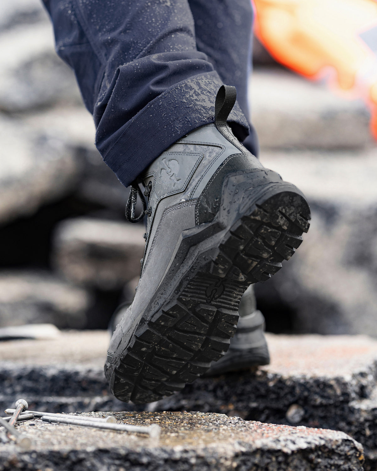 A close-up of a person wearing a pair of rugged black industrial work boots, stepping onto rough concrete blocks outdoors.