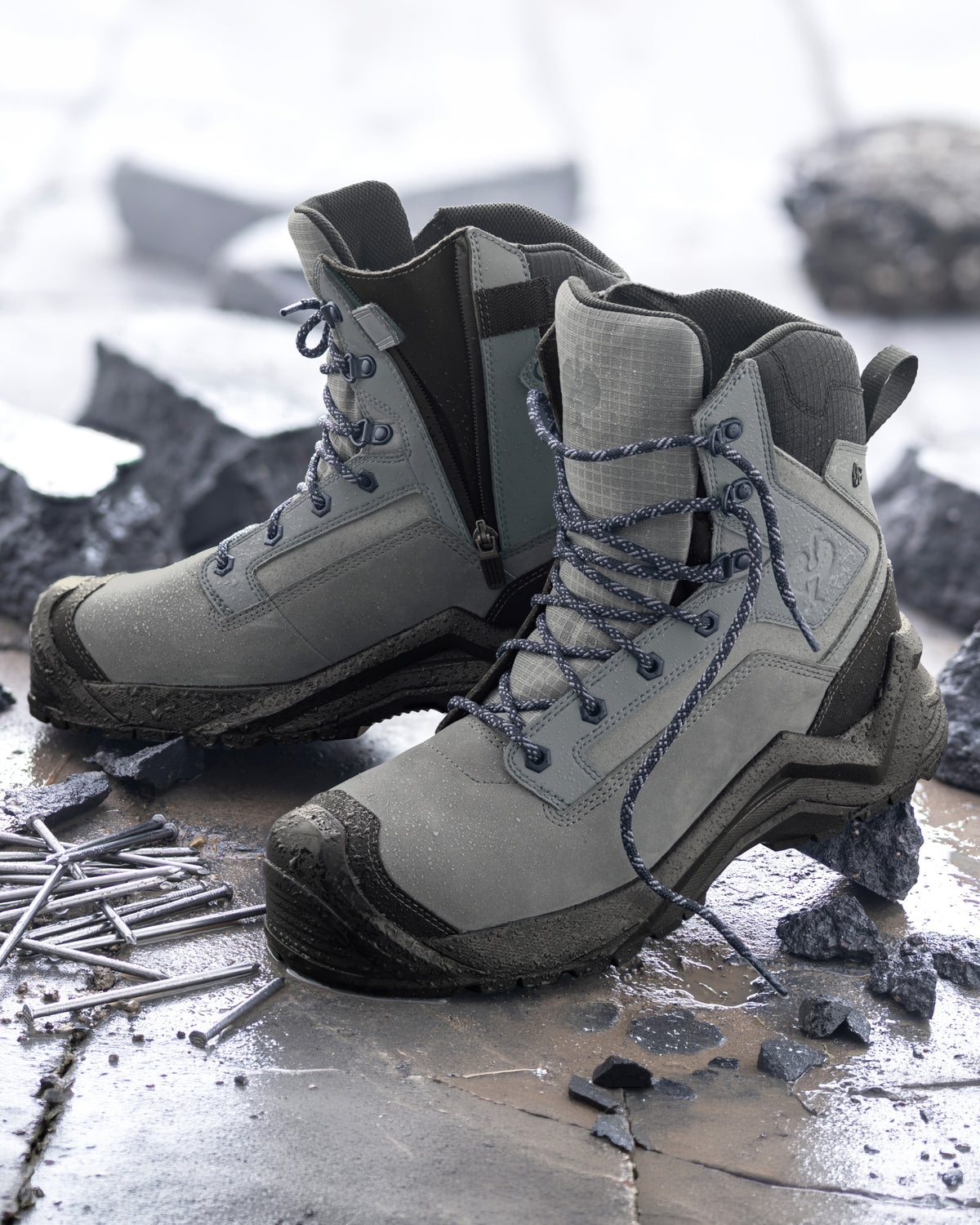 A pair of gray and black rugged lace-up work boots with side zippers, displayed on a wet concrete surface surrounded by rocks and metal nails.