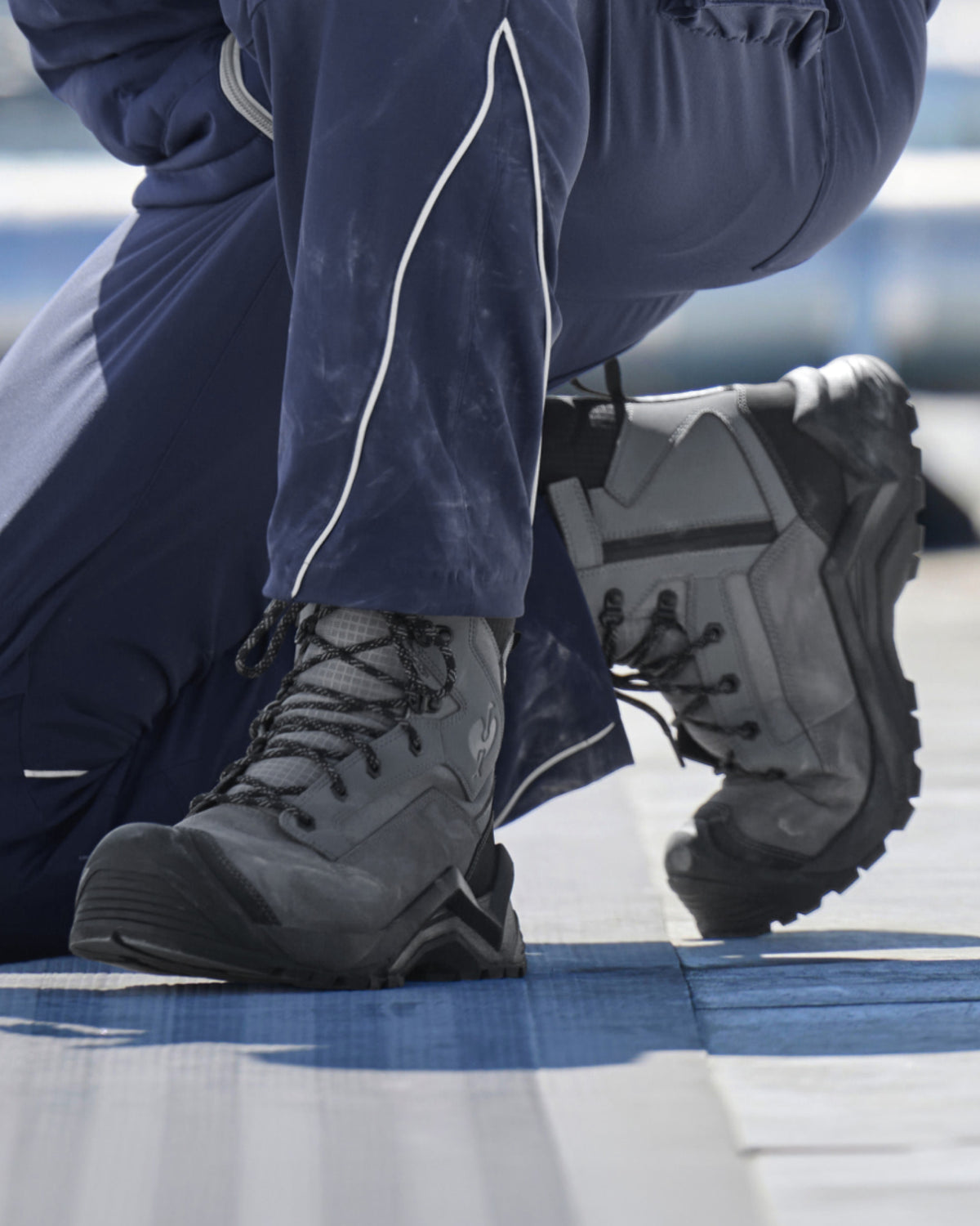 A close-up of a person wearing dark gray and black high-top work boots with navy blue pants, kneeling on a blue and white surface.