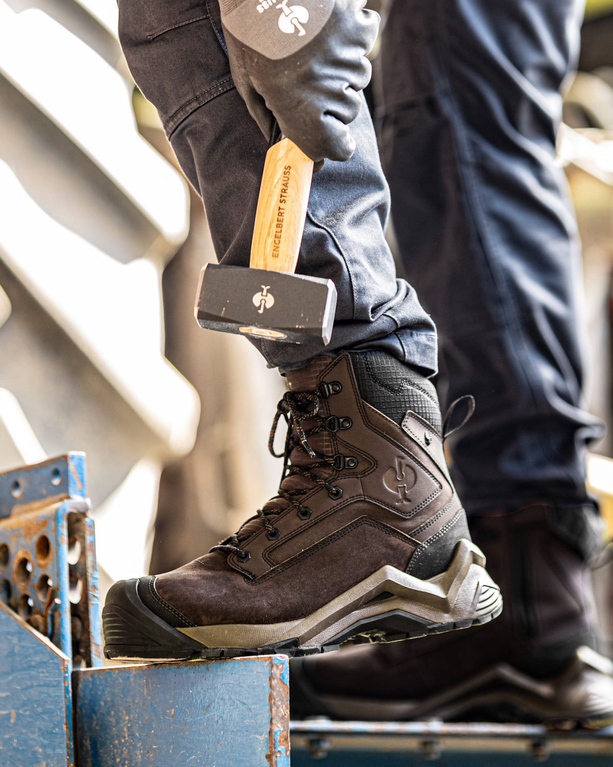 A close-up of a person wearing a dark brown Huk work boot, with their leg raised to demonstrate the pull tab on the heel. They are holding a hammer and wearing dark jeans and a gray work glove in an industrial setting.